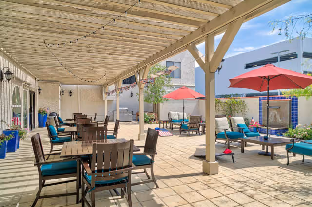 Outdoor patio area with wooden tables and chairs featuring teal cushions under a pergola. Additional seating with teal cushioned chairs and sofas is arranged around tables with red umbrellas. The space is decorated with potted plants and an outdoor water feature is visible on the far wall.