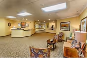 Carpeted reception lobby with a front desk, seating area of chairs and small tables, framed artwork on the walls, and overhead lighting.
