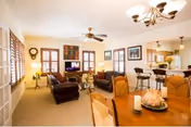A bright and spacious living room area with beige walls and carpet, featuring two dark leather sofas, a wooden coffee table, and a flat-screen TV mounted on the far wall. The room has multiple windows with wooden shutters allowing natural light to enter. A ceiling fan and a chandelier provide lighting. In the foreground, there is a wooden dining table with chairs and a decorative centerpiece. The living room opens into a kitchen area with bar stools along a counter.