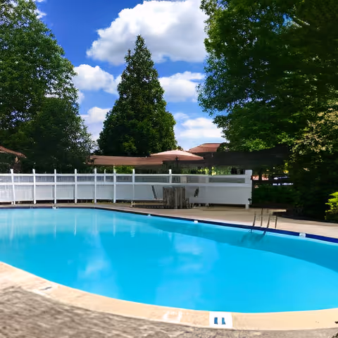 Outdoor swimming pool with clear blue water surrounded by a white fence, trees, and a partly cloudy sky in the background.
