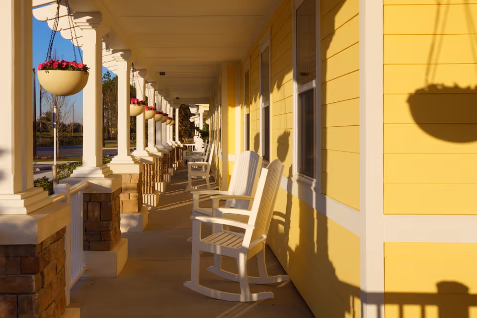 Covered yellow building porch with white rocking chairs and hanging flower baskets along a columned walkway.