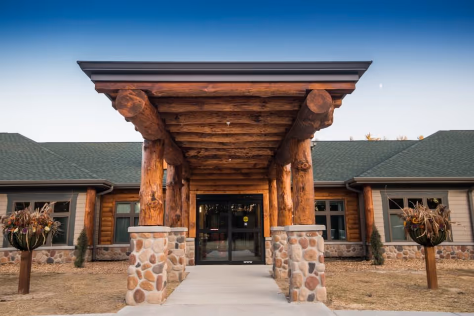 Timber log entrance with stone pillars leads to glass doors under a wooden canopy.