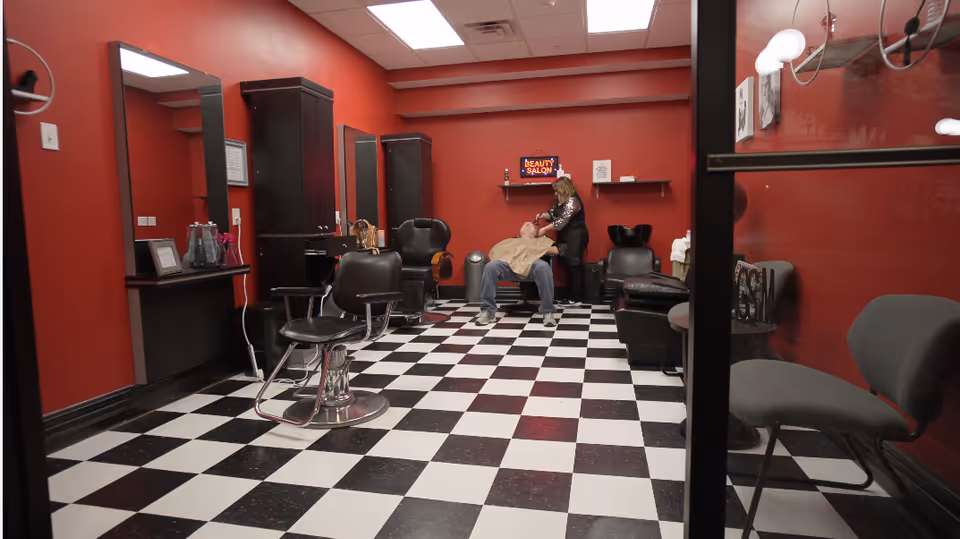 Interior of a beauty salon with red walls and black-and-white checkered floor. A stylist is washing a client's hair in a reclining chair. The room contains salon chairs, mirrors, cabinets, and a neon sign that reads 'BEAUTY SALON'.