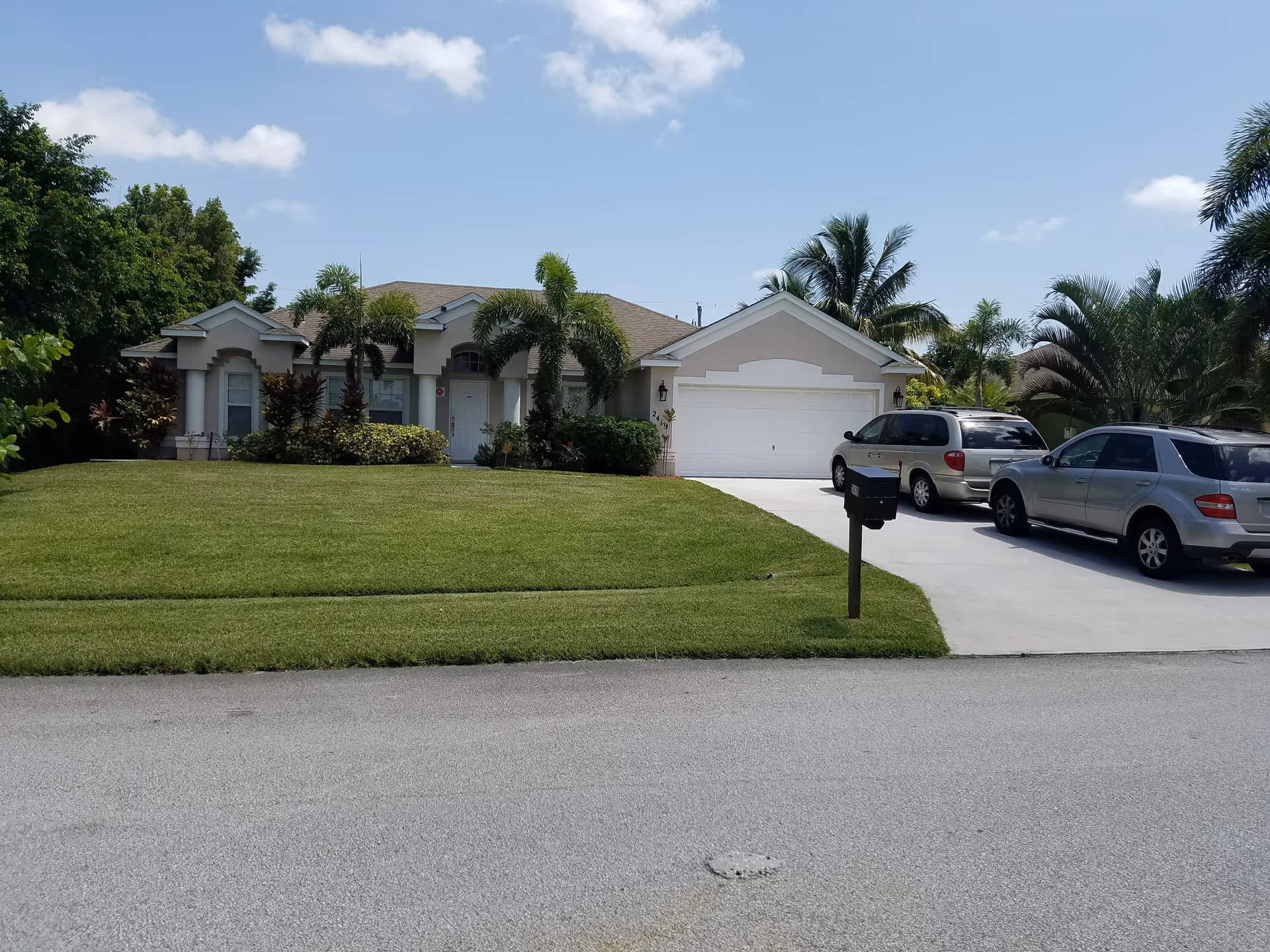 Single-story residential house with a well-maintained front lawn, palm trees, and two cars parked in the driveway under a clear blue sky with a few clouds.