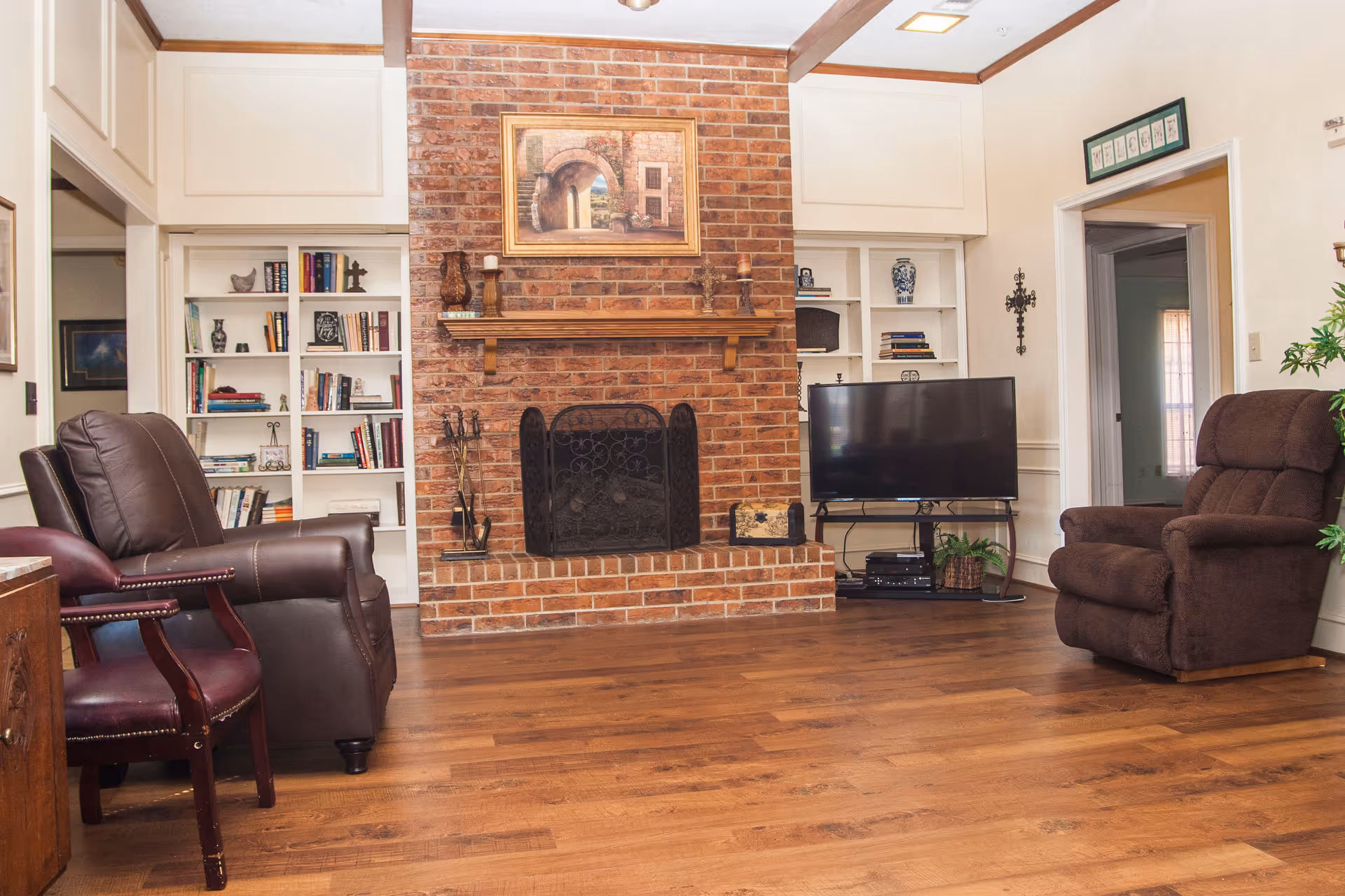 A cozy living room with a brick fireplace in the center, flanked by built-in white bookshelves filled with books and decorative items. There are two brown leather chairs on the left and a brown recliner on the right. A flat-screen TV sits on a stand to the right of the fireplace. The room has wooden flooring and white walls with wooden trim.