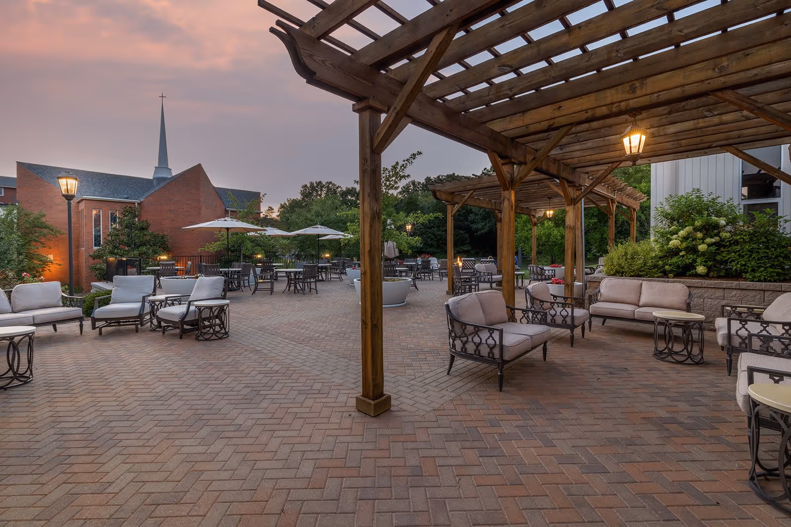 Outdoor patio area at dusk with cushioned seating, metal tables, wooden pergolas with hanging lanterns, and umbrellas. In the background, there is a red brick building with a tall white steeple and surrounding greenery.