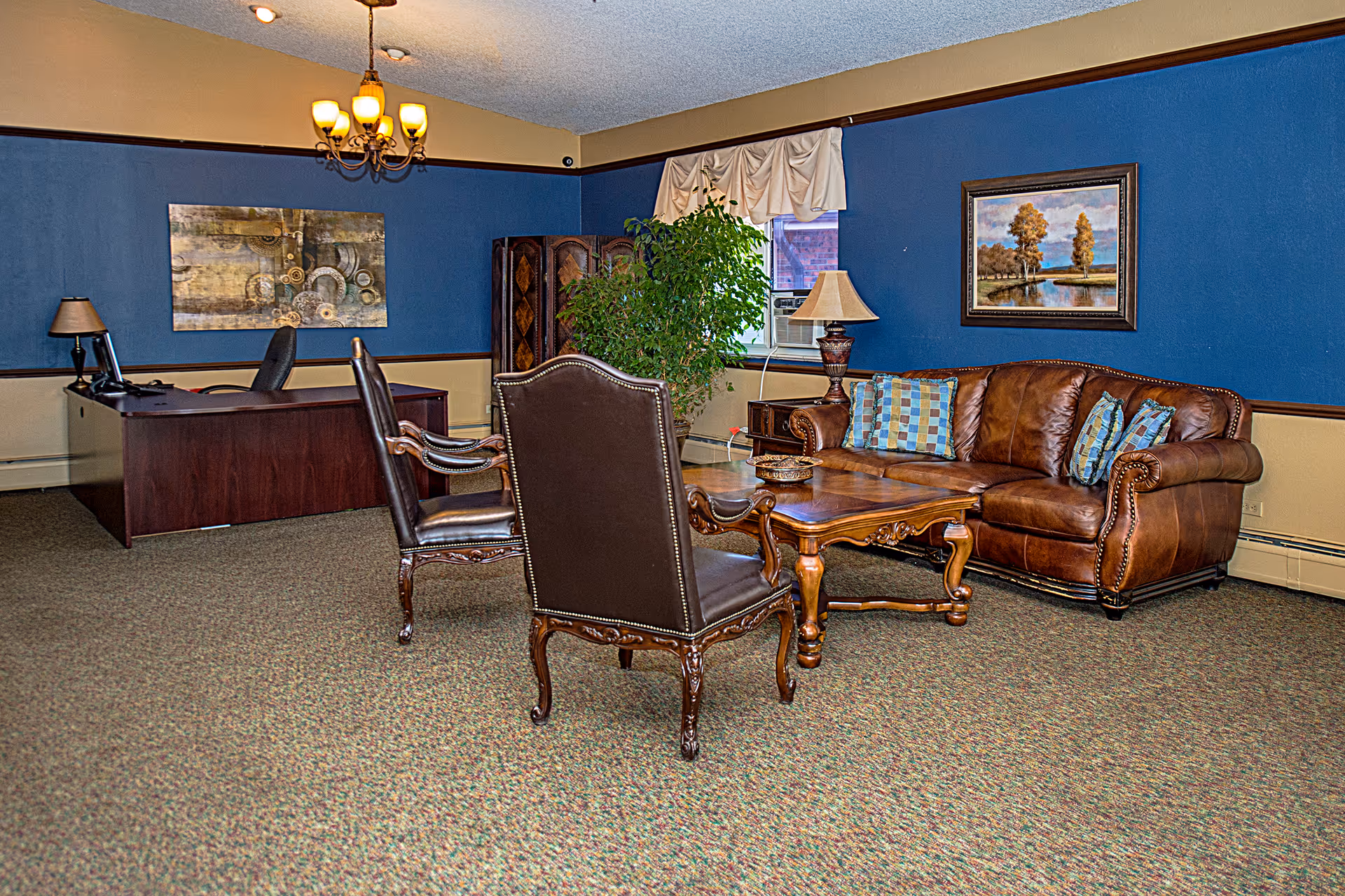 A cozy office and sitting area with a dark wooden desk and chair on the left, and a brown leather sofa with patterned pillows, two dark leather armchairs, and a wooden coffee table on the right. The walls are painted blue with a beige lower section, decorated with framed artwork and a large green plant near a window with a white valance. A chandelier hangs from the ceiling.