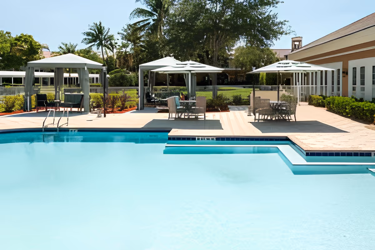 Outdoor swimming pool area with clear blue water, surrounded by a concrete deck. There are three white canopy tents with outdoor seating and tables underneath. Green trees and bushes are visible in the background along with part of a building on the right side.
