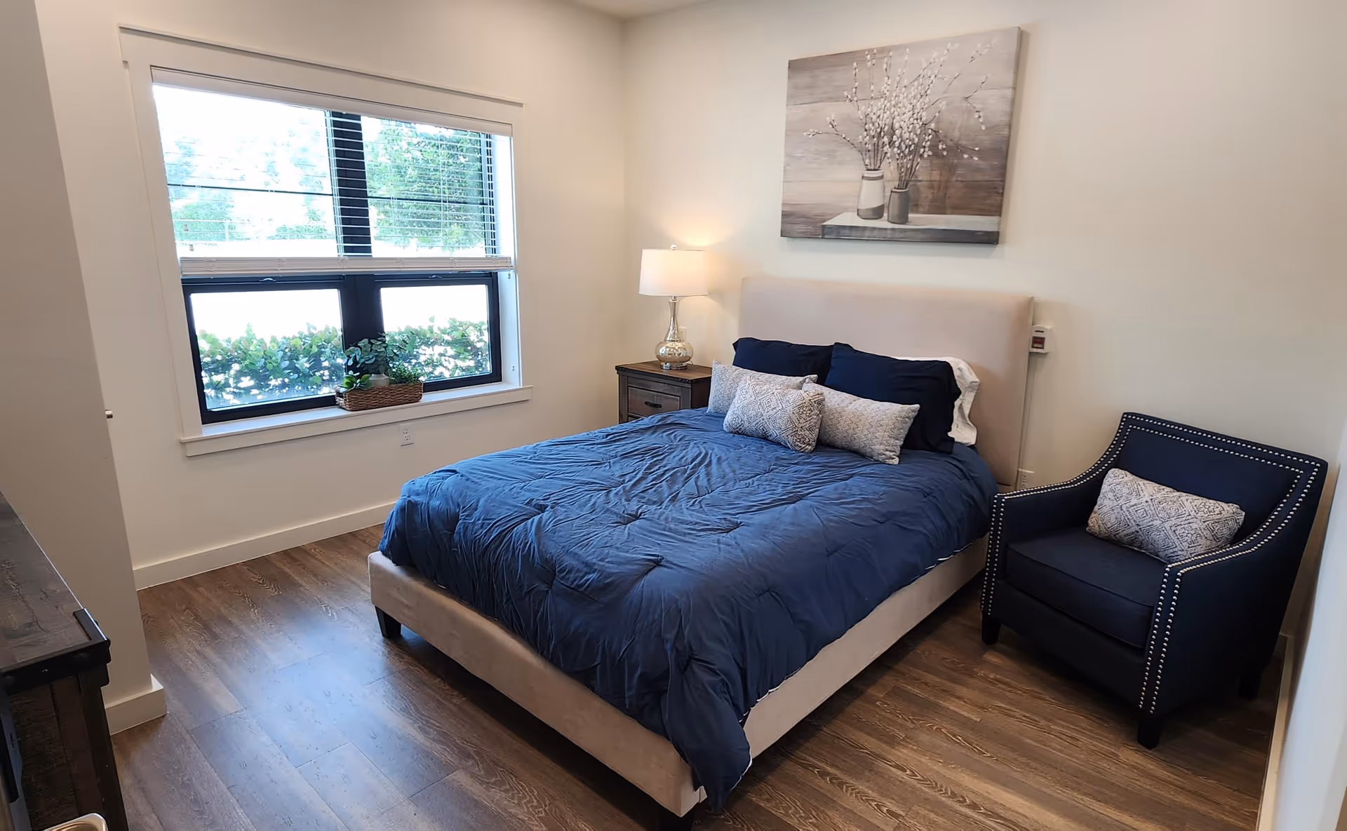 Bright bedroom with a bed dressed in blue bedding, a nightstand and lamp, a navy armchair, and a window showing greenery outside.