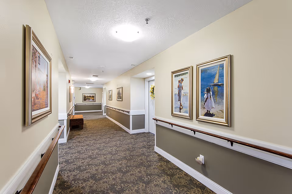 A well-lit hallway in an assisted living facility with beige and olive green walls, handrails on both sides, patterned carpet flooring, framed paintings on the walls, and a bench at the far end.