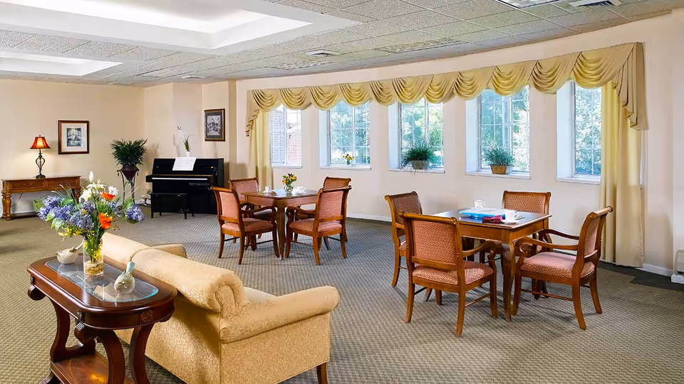 A bright and spacious common area with beige walls and carpeted floor, featuring a beige upholstered sofa, a wooden coffee table with a floral arrangement, two wooden tables each surrounded by four wooden chairs with cushioned seats, large windows with yellow valances letting in natural light, a black upright piano against the far wall, and framed artwork and a lamp on a side table.