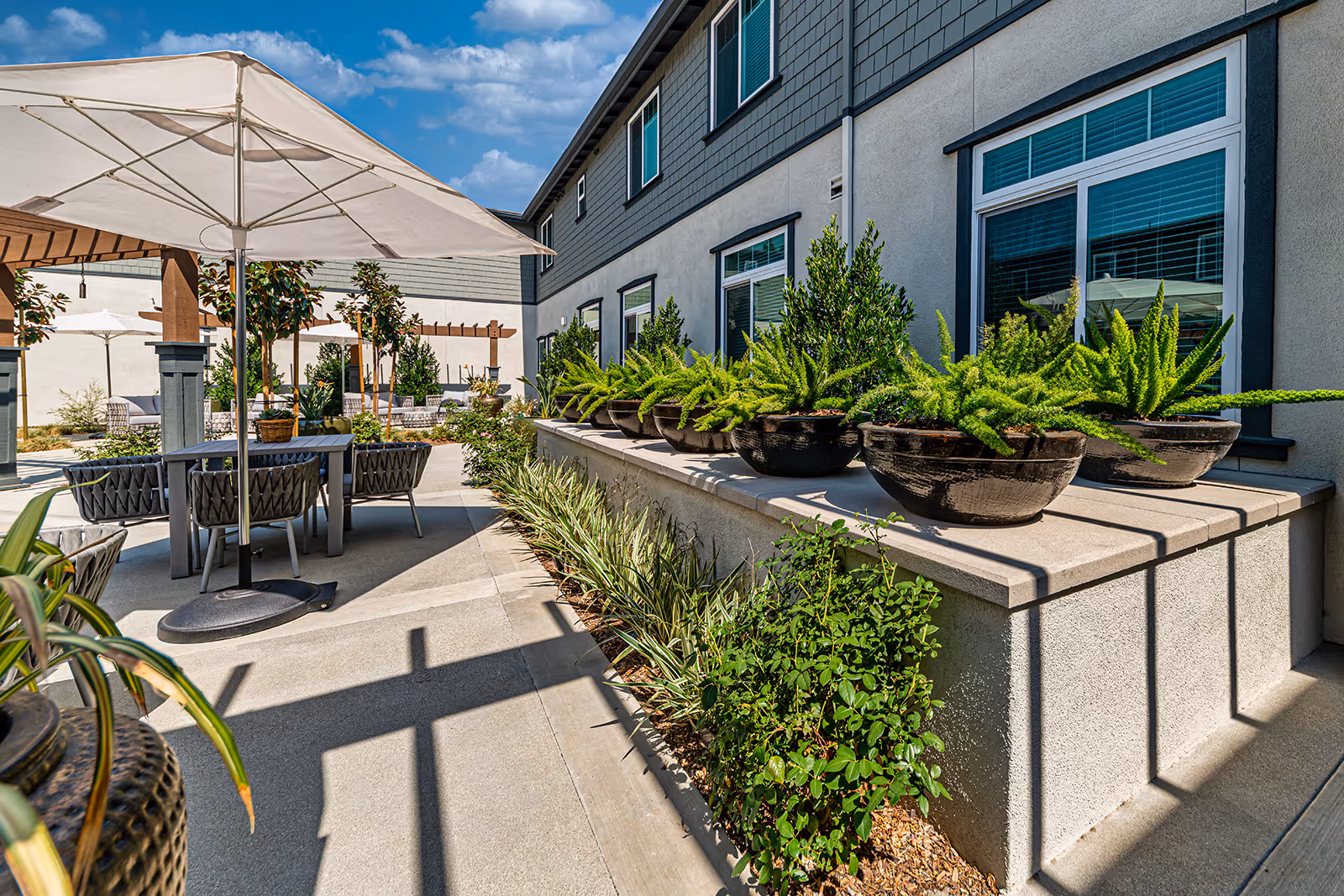 Outdoor patio area at Allara Senior Living featuring tables with umbrellas, chairs, potted plants, and greenery along the building exterior under a blue sky with some clouds.