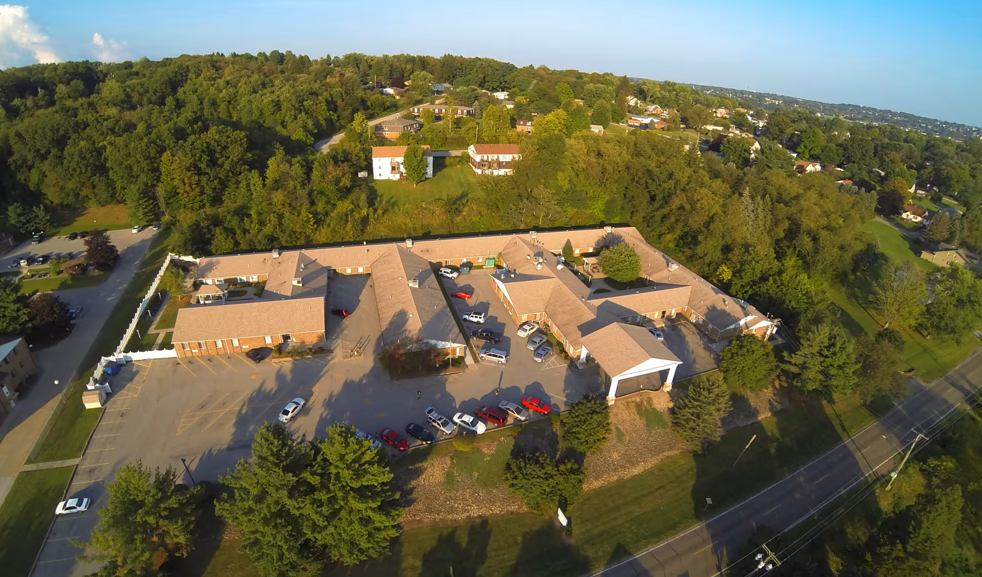 Aerial view of Windsor House at O'Brien Memorial, showing a single-story building with a beige roof surrounded by trees and greenery. Several cars are parked in the parking lot adjacent to the building, and a road runs along the right side of the image.