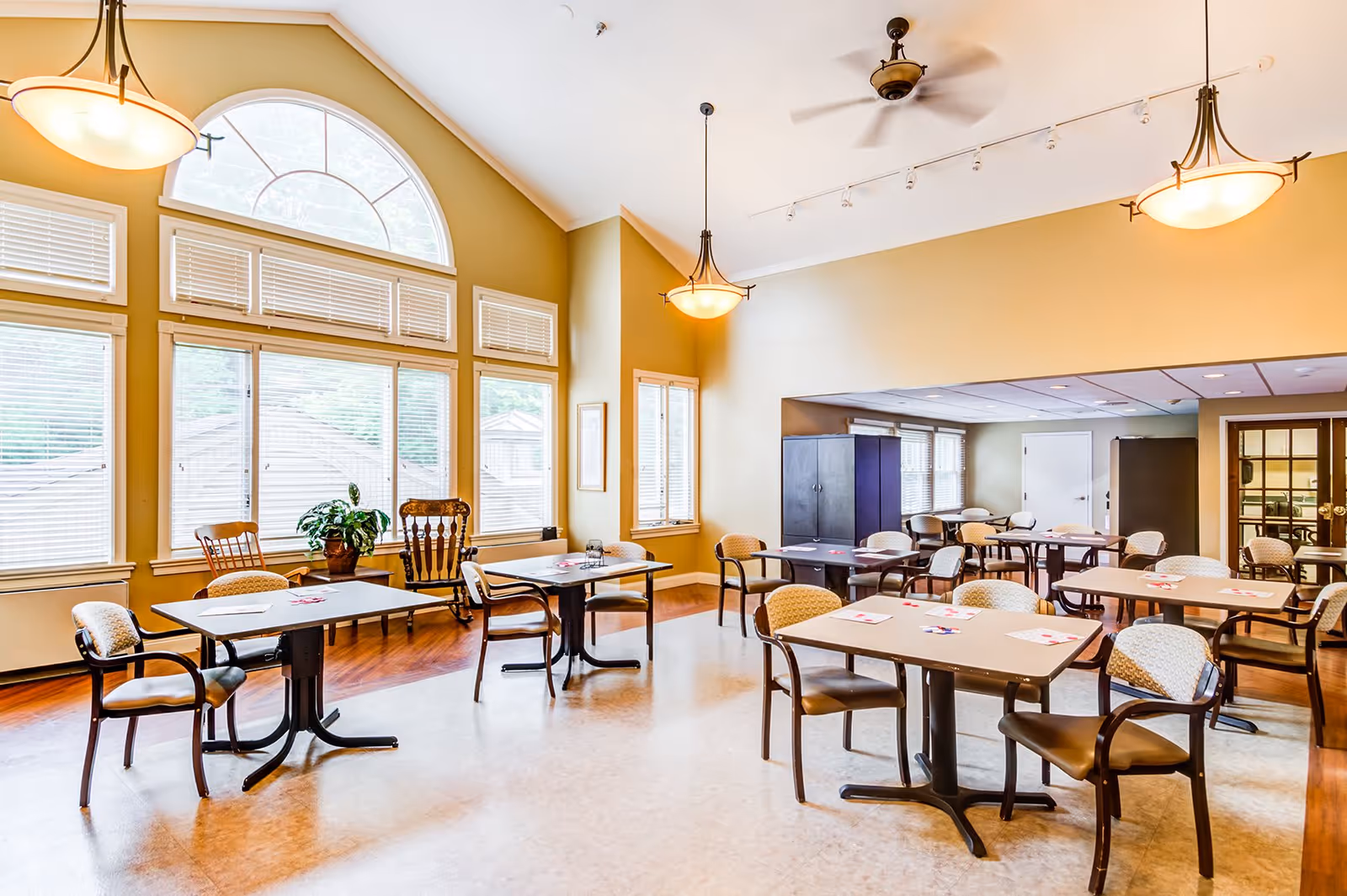 A bright dining room with multiple tables and chairs arranged neatly. Large windows with blinds allow natural light to fill the room. The walls are painted a warm beige color, and there are hanging light fixtures from the ceiling. A potted plant and a wooden chair are placed near the windows.