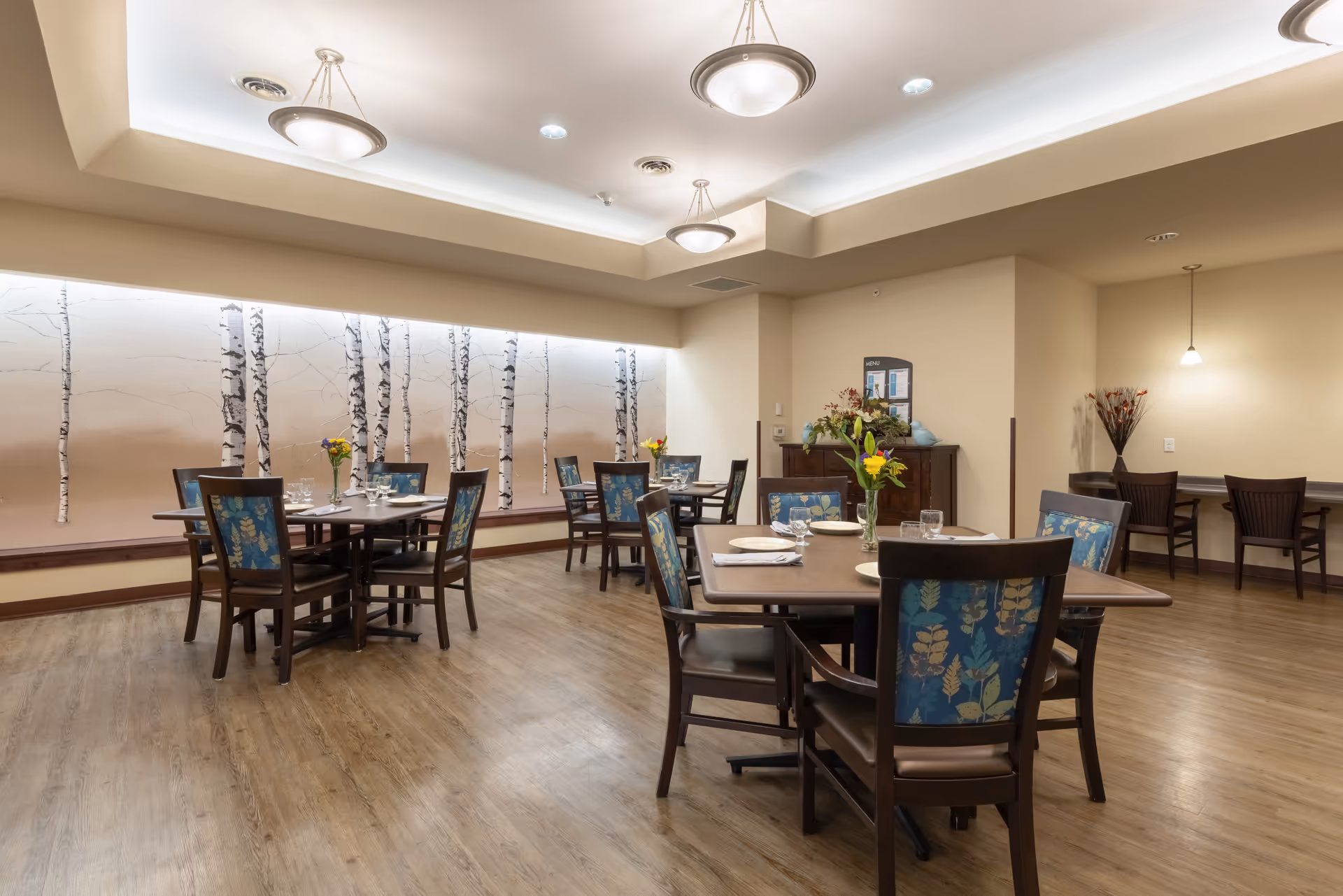 Spacious dining room with multiple set tables and patterned chairs, a birch-tree mural on the wall, and soft overhead lighting.