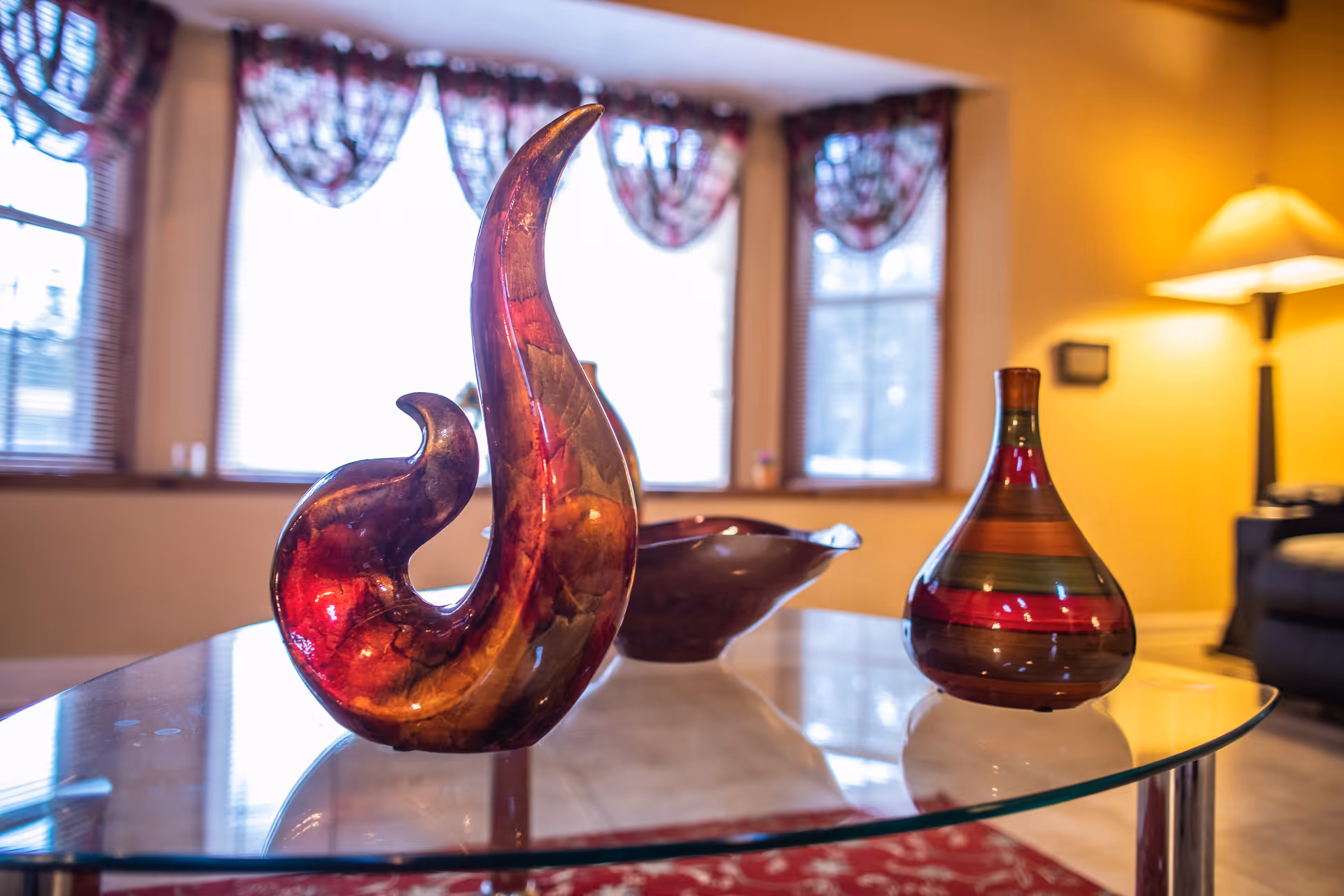 A close-up view of decorative glass art pieces on a glass coffee table in a warmly lit living room with windows covered by floral valances and a floor lamp in the background.