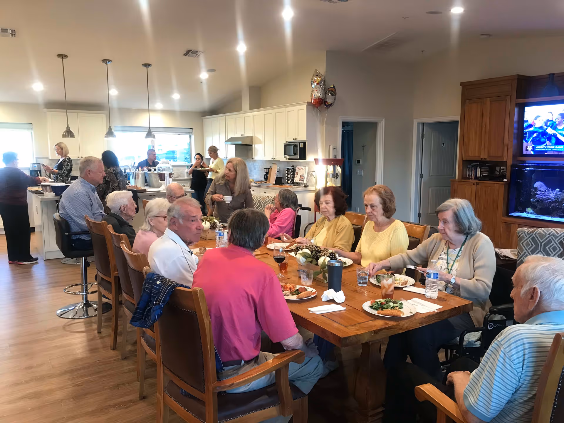 A group of elderly people sitting around a wooden dining table eating a meal in a well-lit kitchen and dining area. Several people are seated at the table with plates of food and drinks, while others are standing in the kitchen area serving themselves. The room has white cabinets, pendant lights, and a television mounted on a wooden cabinet with an aquarium below it.