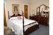 Traditional bedroom with a wooden four-poster bed, matching dresser with mirror, armchair, and framed artwork on the wall.