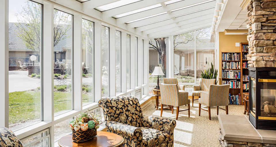 A bright and cozy living room area with large floor-to-ceiling windows letting in natural light. The room features a stone fireplace on the right, a bookshelf filled with books, a round wooden table with a decorative centerpiece, and several upholstered chairs arranged for conversation. Outside the windows, a garden and patio area are visible.