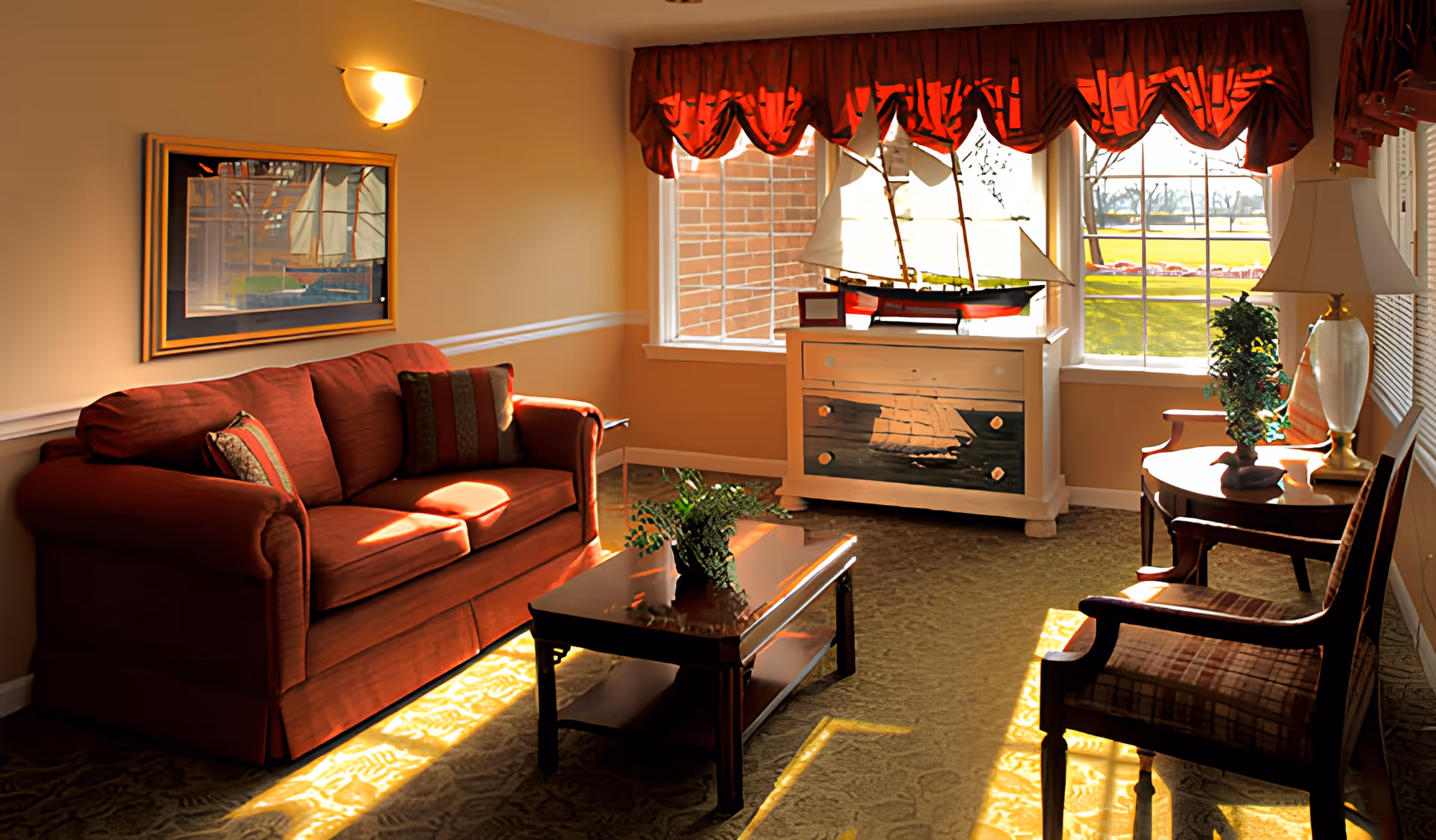 Sunlit living room with a red sofa, coffee table, armchairs, and a decorative ship on a chest beneath a window with a red valance.