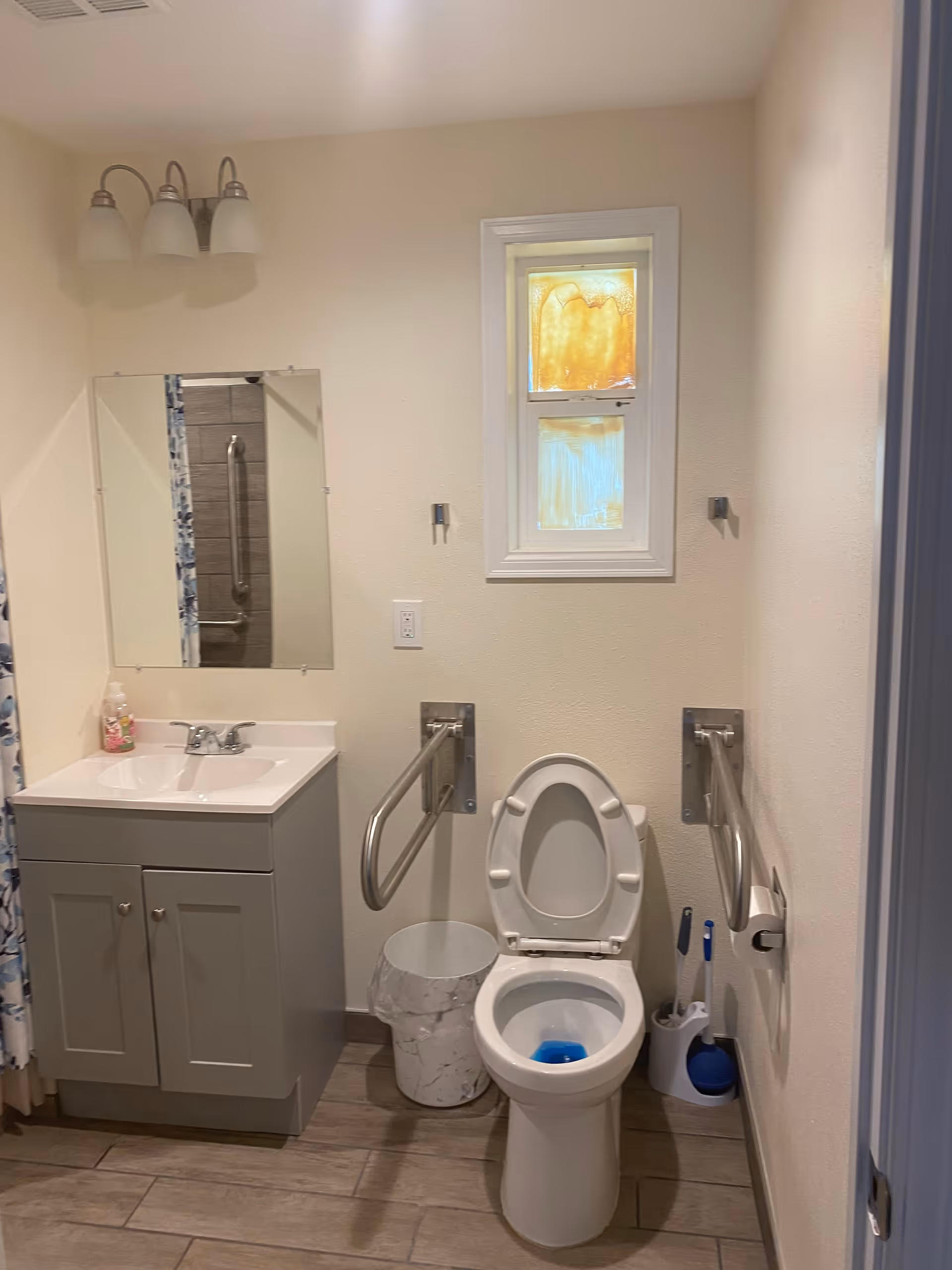 Small accessible bathroom with a toilet (open lid), wall-mounted grab bars, a sink vanity with mirror, a small window, and tiled floor.