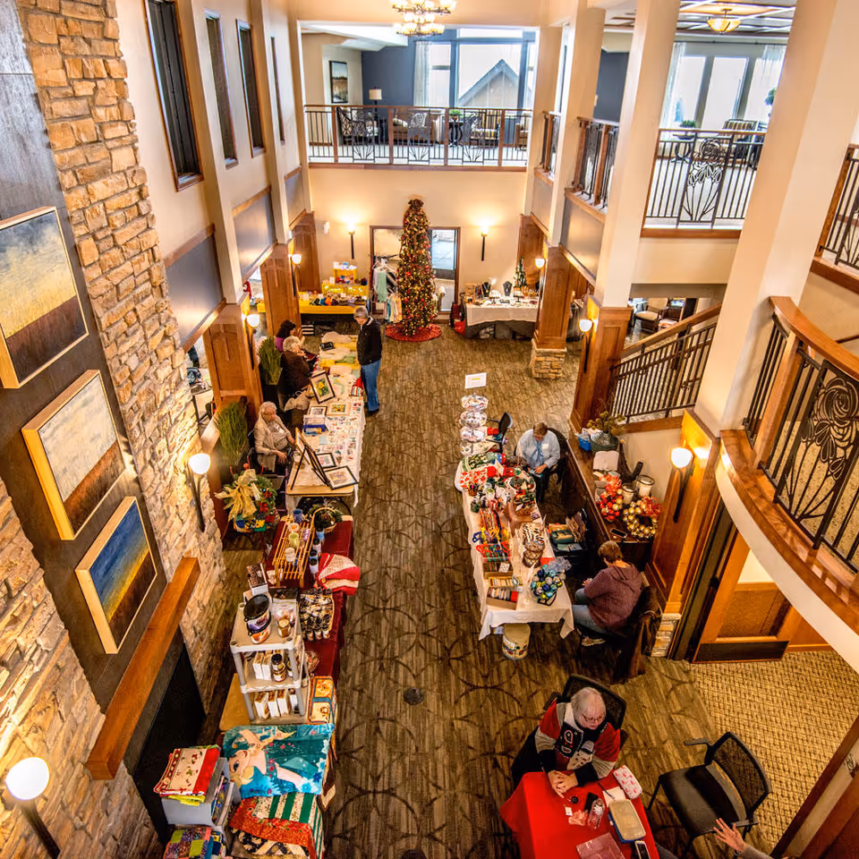 Interior view of a senior living facility lobby decorated for the holidays with a Christmas tree in the center. Several tables are set up along the sides with people sitting and standing behind them, displaying various items for sale or exhibition. The space has a high ceiling, stone wall with framed artwork, and a staircase with decorative railings.