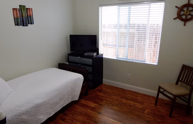 A small bedroom with a single bed covered in a white quilt, a black dresser with a TV on top, a wooden chair with a cushion, a window with blinds, and wall decorations including a colorful abstract piece and a ship's wheel.