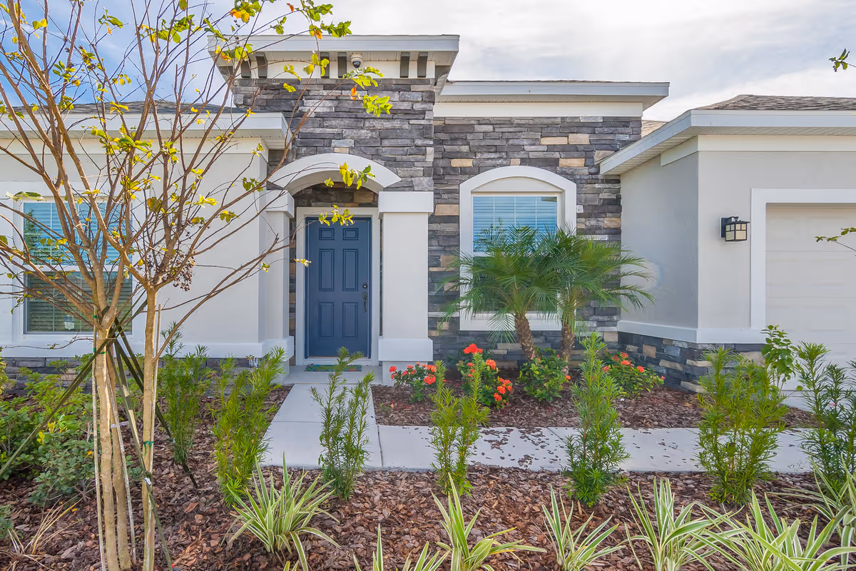Front exterior view of a single-story house with a blue front door, stone and stucco facade, a window with white blinds, and landscaped garden with small palm trees, flowering plants, and shrubs.