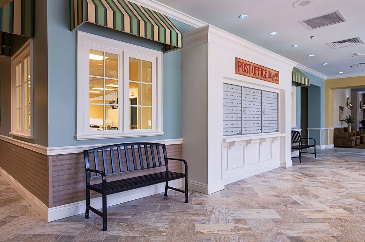 Interior hallway of a senior living facility with a black bench in front of a blue wall featuring windows with striped green and beige awnings. On the right side, there is a white wall with multiple mailboxes labeled 'POST OFFICE DELAND FL 32720'. The floor is tiled with a beige and brown pattern, and there are additional benches and seating areas visible further down the hallway.