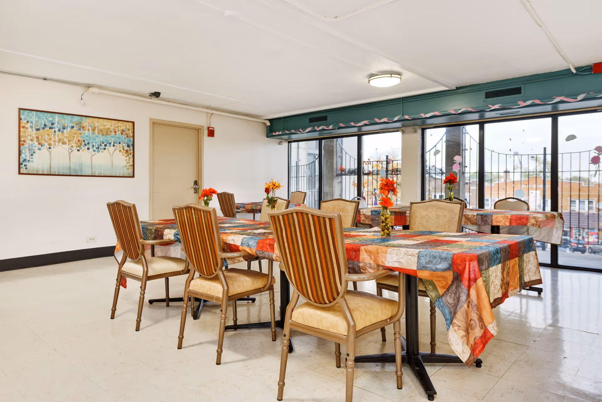 A dining room with several tables covered in colorful patchwork tablecloths, each adorned with a vase of red and orange flowers. The room has large windows with a view of the outside, beige walls, and a framed abstract painting on one wall. There are multiple chairs with striped upholstery around the tables.