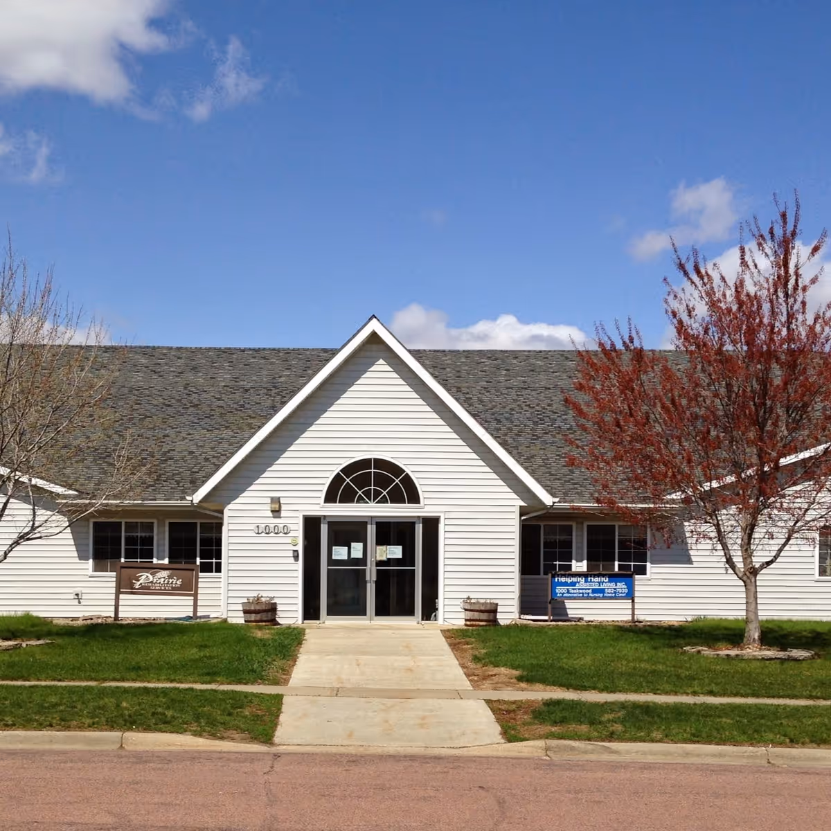 Front exterior view of a single-story building with white siding and a gray shingled roof under a blue sky with some clouds. There is a concrete walkway leading to double glass doors with an arched window above. Two trees flank the entrance, one with red leaves and the other bare. Signs are visible near the entrance, including one that reads 'Helping Hand Assisted Living'.