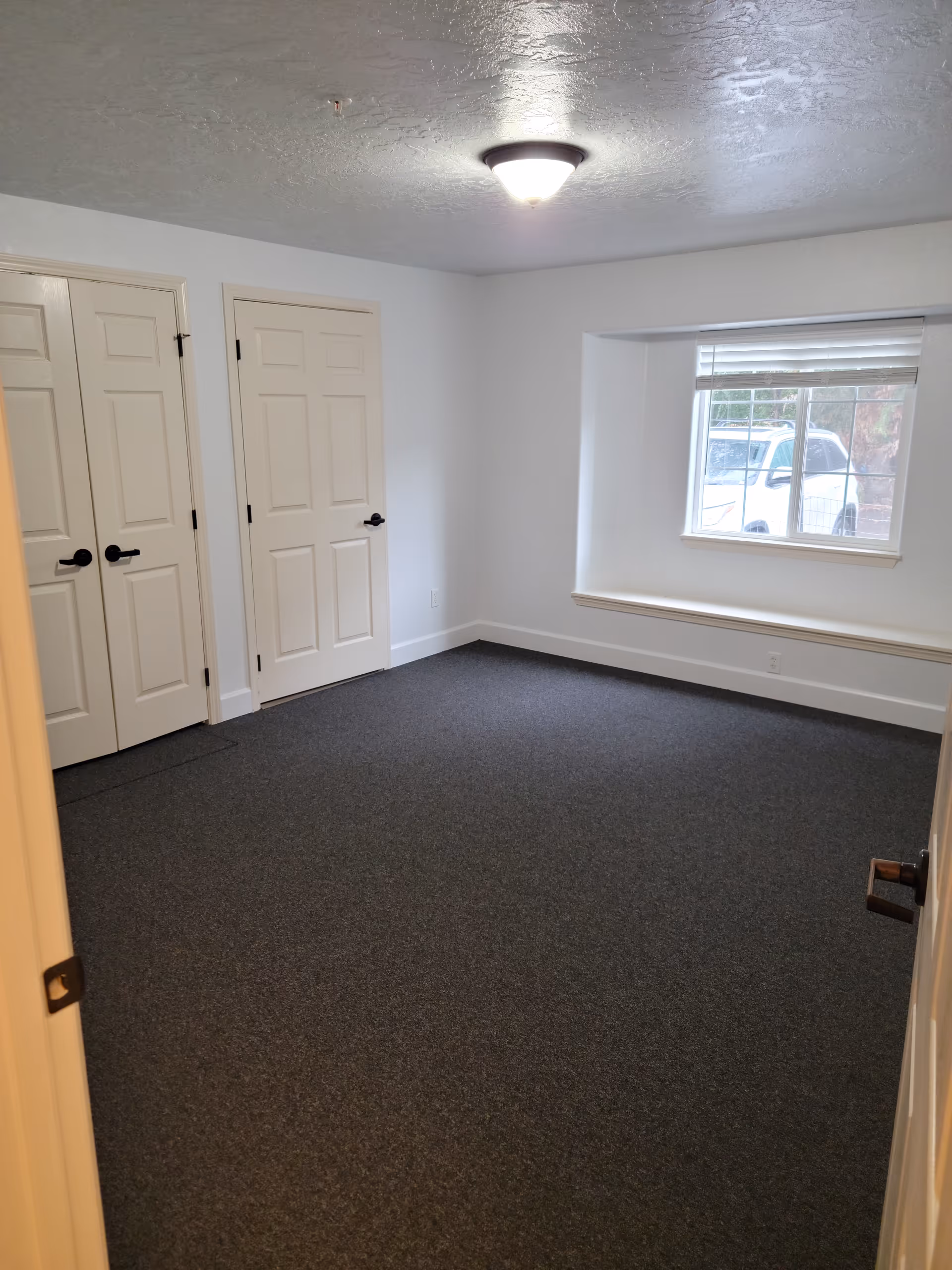 Empty carpeted bedroom with white walls, two closet doors, a window seat, and an overhead light.