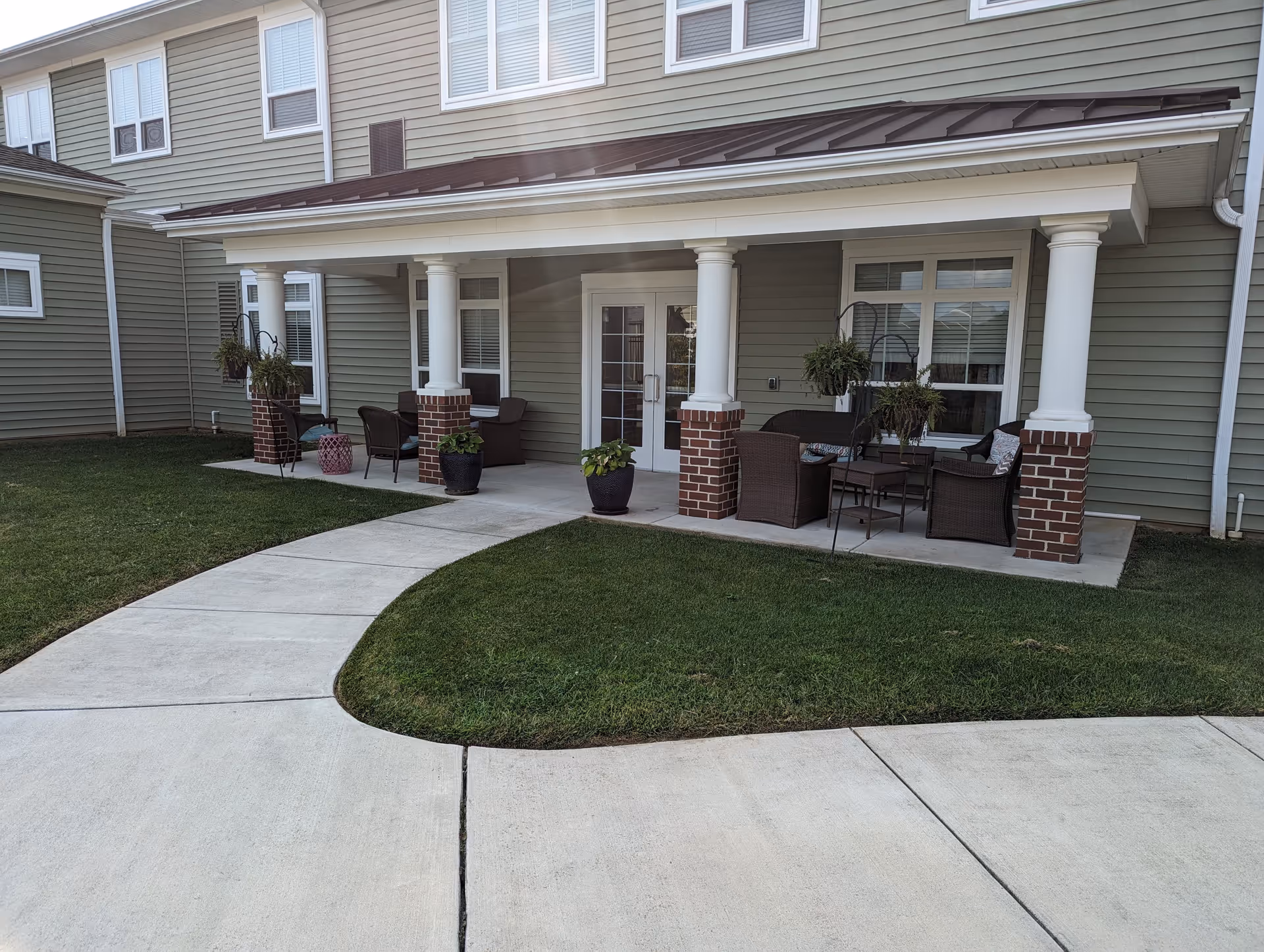Outdoor patio area of a senior living facility with a covered porch supported by white columns with brick bases. The porch has several wicker chairs and small tables, along with potted plants and hanging planters. A curved concrete walkway leads up to the porch, surrounded by well-maintained green grass.