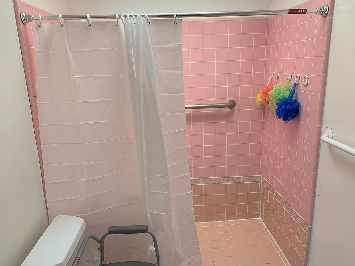 Bathroom with pink tiled walls and floor, a white shower curtain partially drawn, a metal grab bar on the wall, three colorful bath poufs hanging on hooks, and a white toilet with a gray walker in front of it.