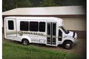 A white shuttle bus parked on grass in front of a beige metal building. The bus has the text 'Sena Kean Manor' on its side along with a logo.