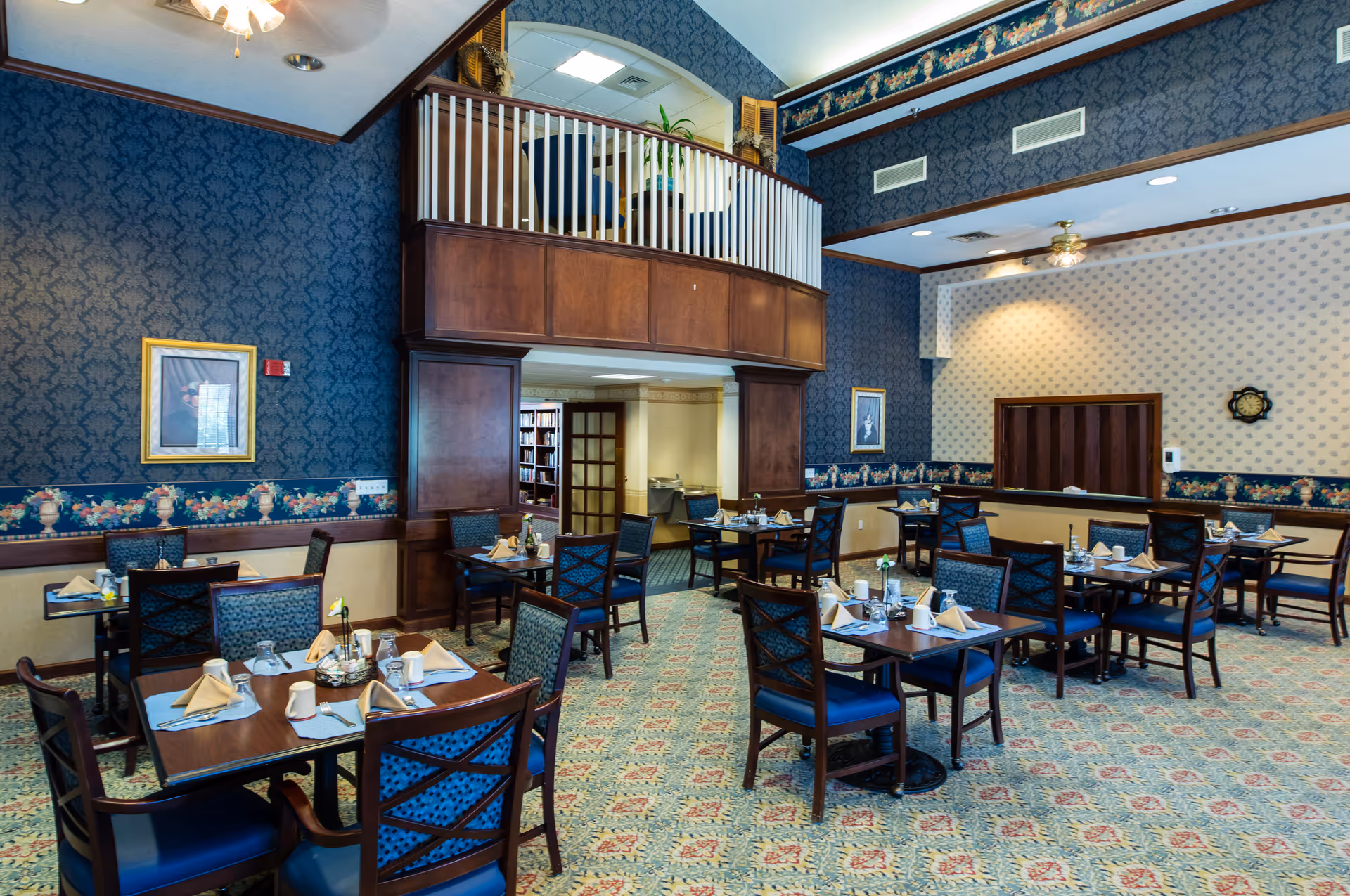 A dining room in an assisted living facility with multiple tables set for meals. Each table has chairs with blue cushions, napkins, cups, and condiments. The room features patterned wallpaper with floral borders, a carpeted floor with a geometric design, and a ceiling fan. There is a wooden balcony railing above a doorway leading to another room.