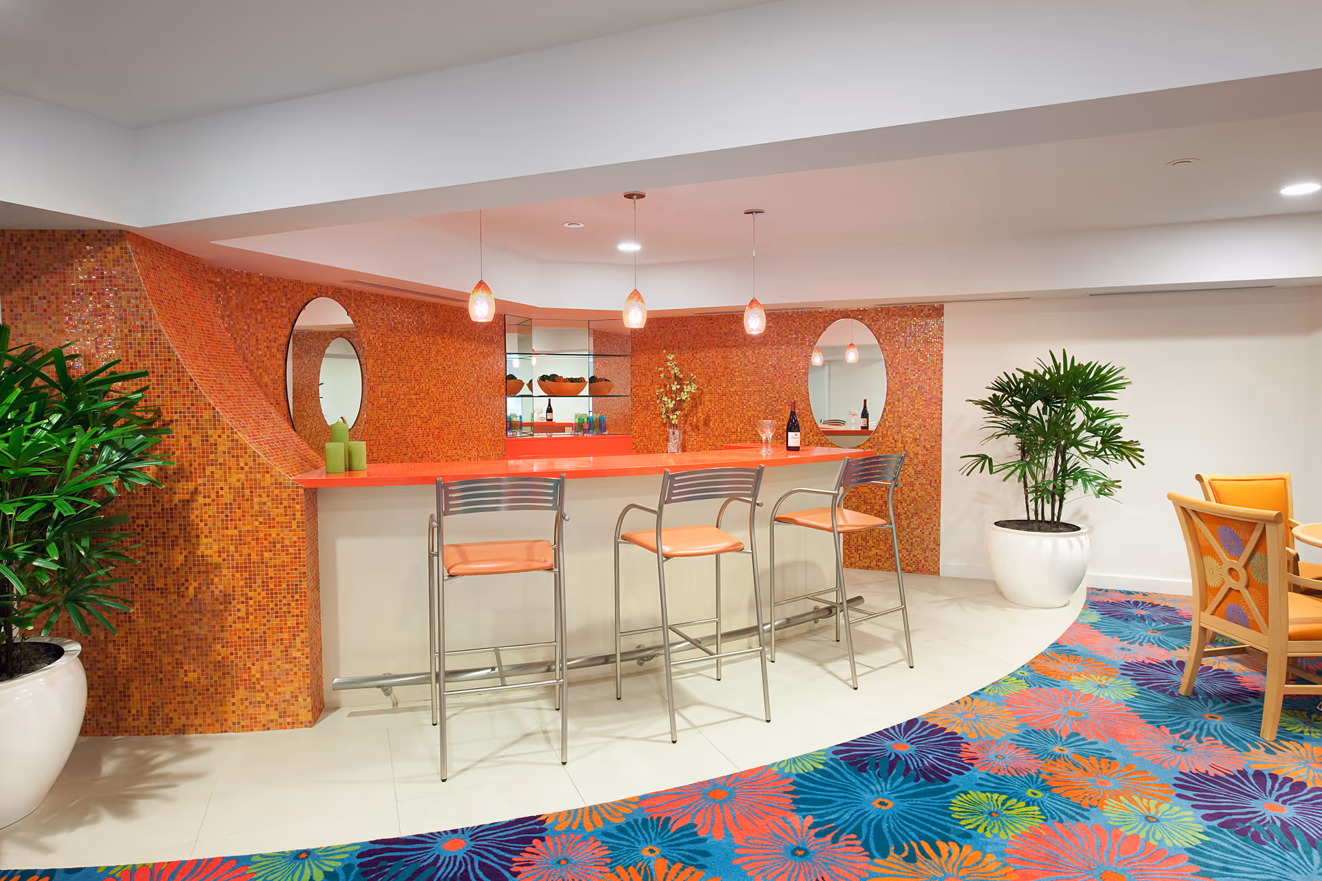 A brightly colored indoor bar area with an orange mosaic tile wall and countertop. There are three metal bar stools with orange seats in front of the bar. Two oval mirrors are mounted on the wall behind the bar, reflecting the pendant lights hanging above. The floor features a vibrant floral patterned carpet in blue, orange, green, and purple hues. Two large potted plants flank the bar area, and a small dining table with orange cushioned chairs is partially visible on the right.