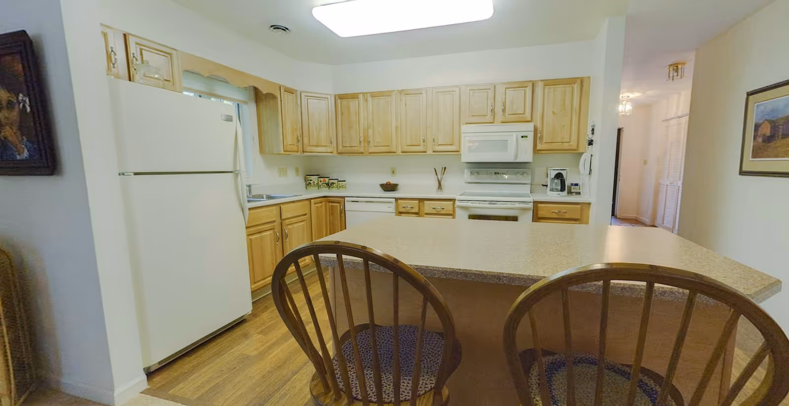 A kitchen with wooden cabinets, a white refrigerator, a white microwave above a white stove, a dishwasher, and a countertop island with two wooden chairs. The floor is wooden, and there are framed pictures on the walls.