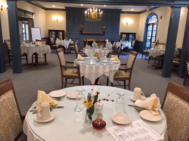 A dining room with multiple round tables covered with white tablecloths, each set with plates, glasses, napkins, and silverware. The room features a chandelier, wall sconces, a fireplace with decorative items on the mantel, and large windows with arched tops. Chairs with patterned upholstery surround the tables.