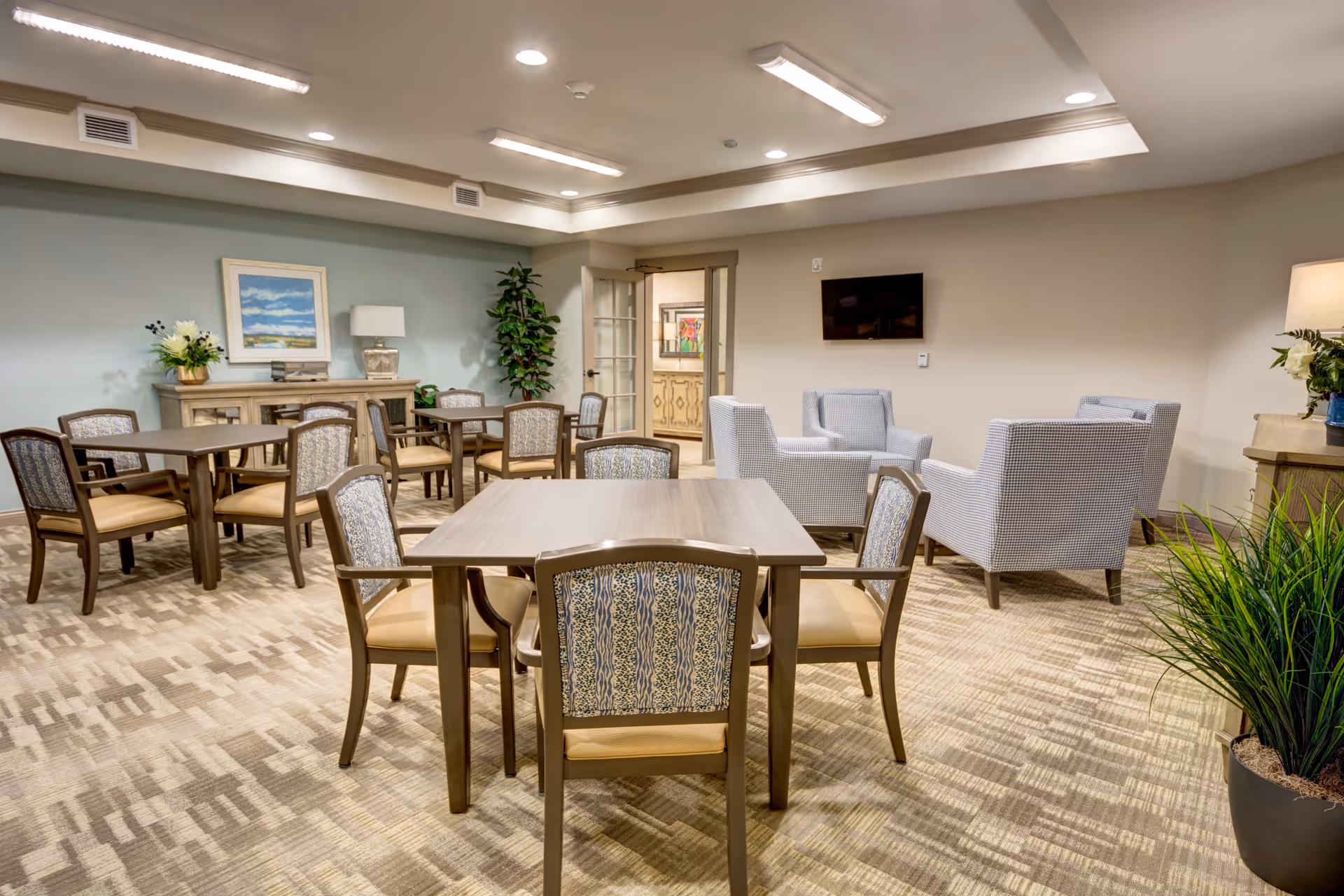 A well-lit common area in a senior living facility featuring several wooden tables with cushioned chairs arranged around them. In the background, there are four upholstered armchairs positioned in a conversational grouping near a wall-mounted flat-screen TV. The room is decorated with plants, a table lamp, and framed artwork on the walls. The carpet has a patterned design, and the ceiling has recessed lighting.