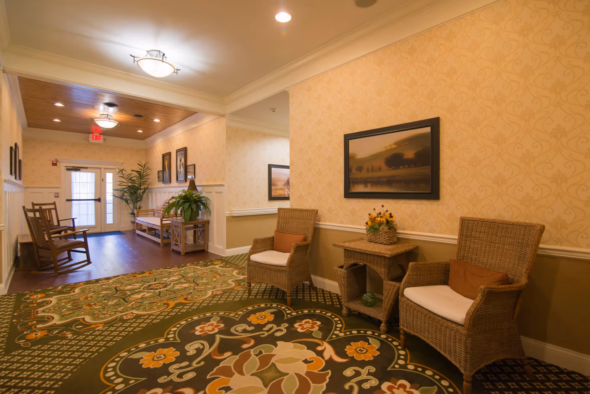 Warmly lit assisted living sitting area with wicker chairs, a side table with flowers, patterned carpet, and a hallway leading to an exit door.