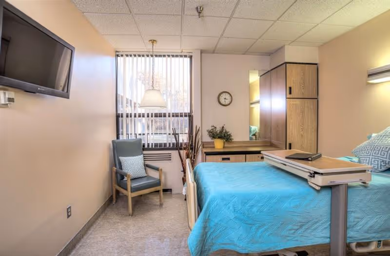 A small, neatly arranged bedroom in a rehabilitation center featuring a hospital bed with a blue bedspread, an adjustable overbed table, a wooden chair with a cushion, a wall-mounted flat screen TV, a window with vertical blinds, a wooden dresser with a potted plant, and a wall clock.