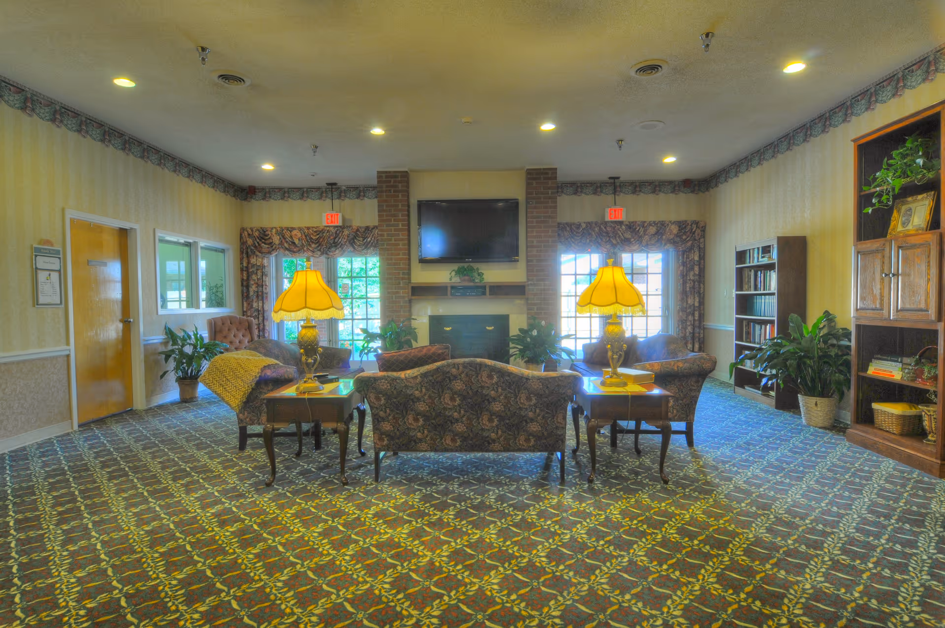 A cozy living room area in a senior living facility with patterned carpet and wallpaper. The room features a central fireplace with a mounted flat-screen TV above it, flanked by two large windows with floral curtains. There are two yellow lamps on side tables beside floral upholstered sofas and armchairs arranged around the fireplace. Bookshelves and potted plants are placed along the walls, creating a warm and inviting atmosphere.