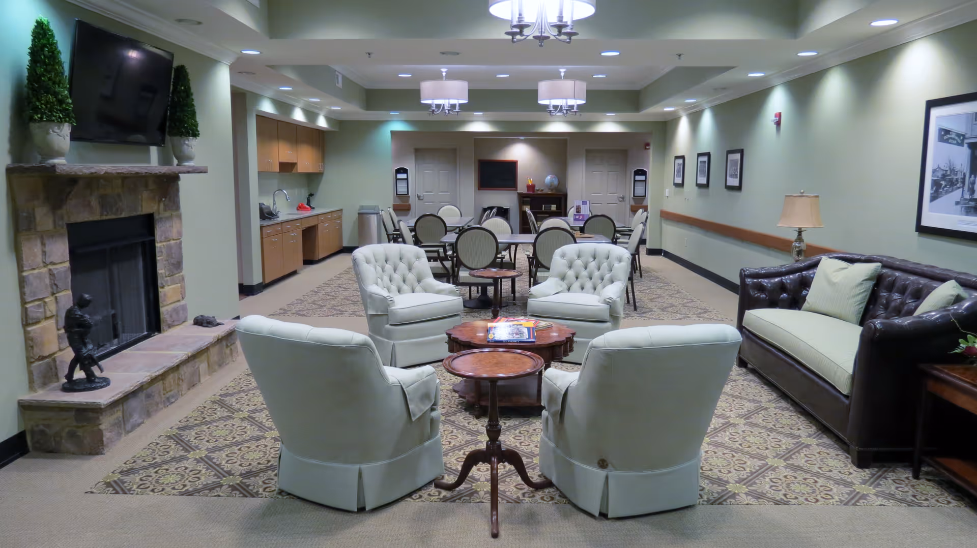 A cozy senior living community common area with a stone fireplace, a wall-mounted TV, four cushioned armchairs arranged around a set of small wooden tables, a dark leather sofa with light cushions, and a kitchenette area with cabinets and a sink. The room has soft green walls, framed pictures, and overhead lighting fixtures.