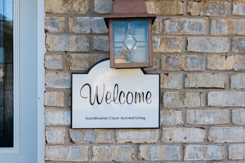 A white welcome sign with black text that reads 'Welcome Scandinavian Court Assisted Living' mounted on a light gray brick wall beneath a decorative outdoor lantern.