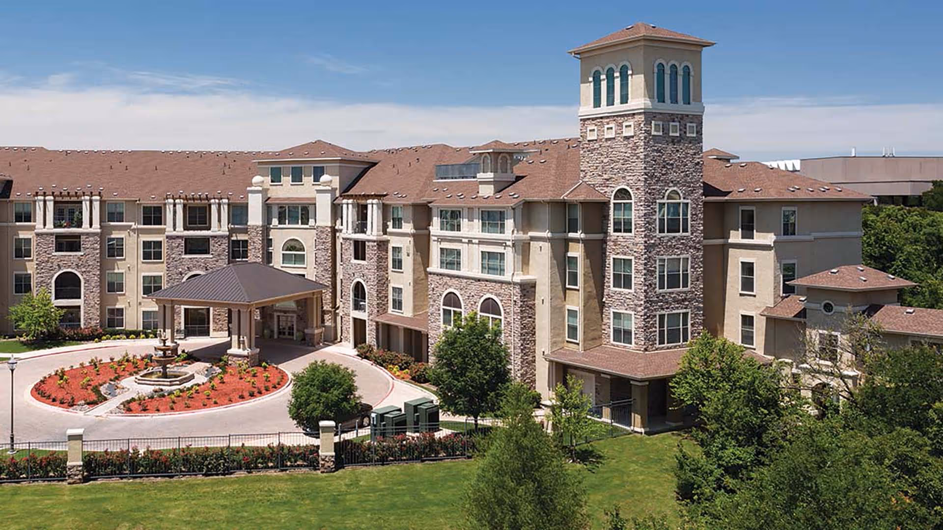 Exterior view of a large senior living facility building with stone and beige walls, multiple windows, a covered entrance, and a landscaped circular driveway with a fountain in the center. The building has a tower-like structure on the right side and is surrounded by green lawns and trees under a blue sky.