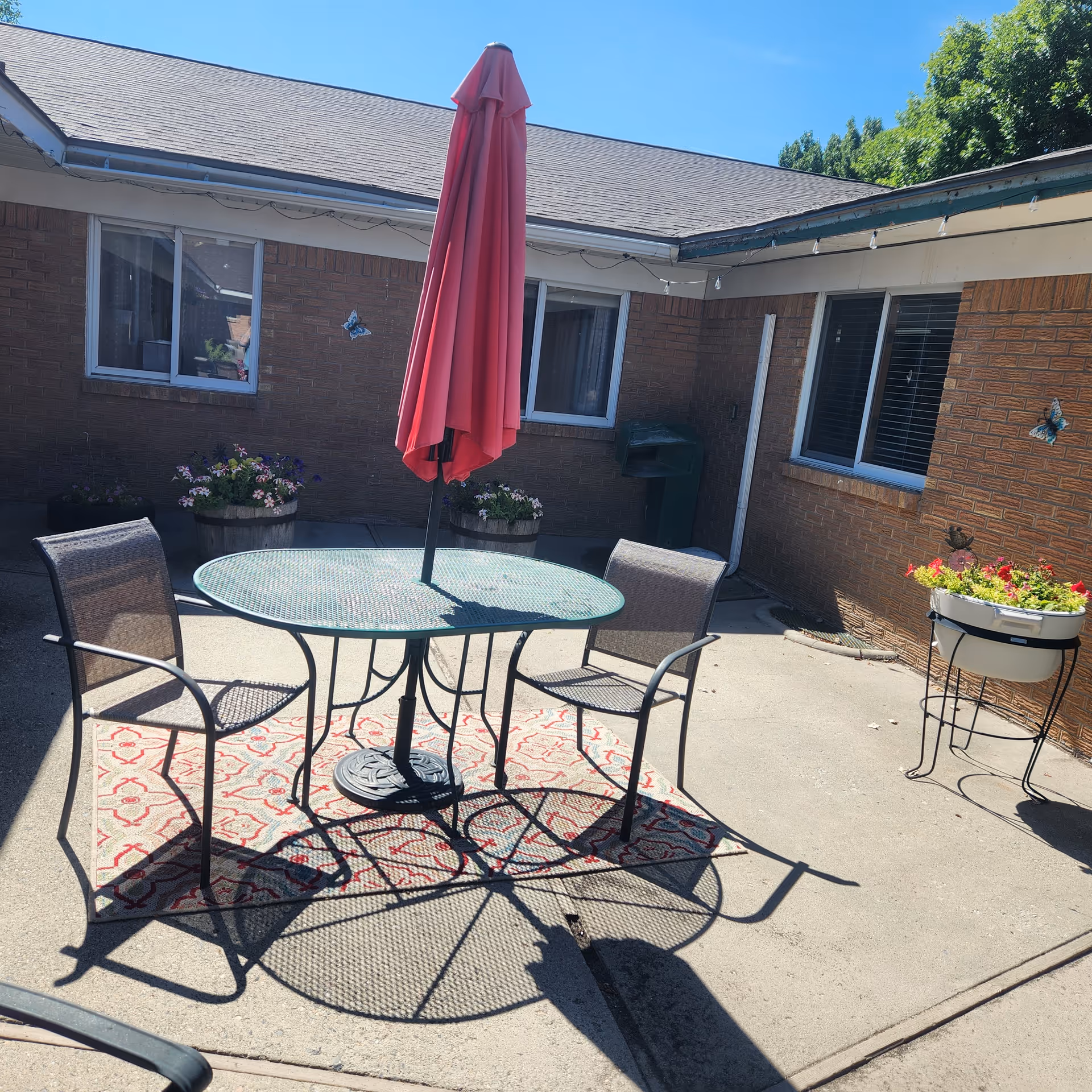 Outdoor patio area with a round metal table and two metal chairs on a patterned outdoor rug. A closed red umbrella is positioned in the center of the table. The patio is surrounded by brick walls with windows, flower pots with colorful flowers, and some butterfly decorations on the walls. The sky is clear and blue.