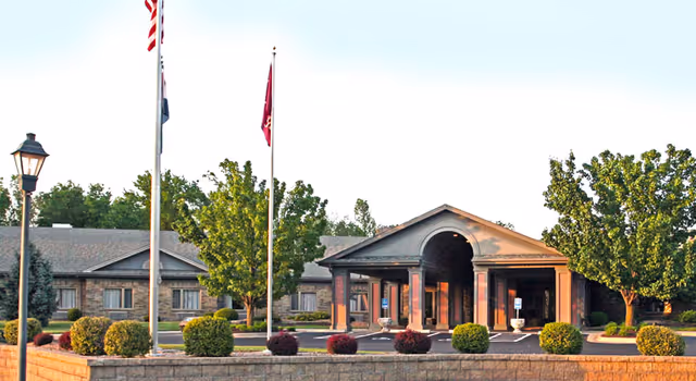 Front exterior view of Manor At Elfindale facility showing a single-story building with a covered entrance, surrounded by trees, bushes, and flagpoles with flags.