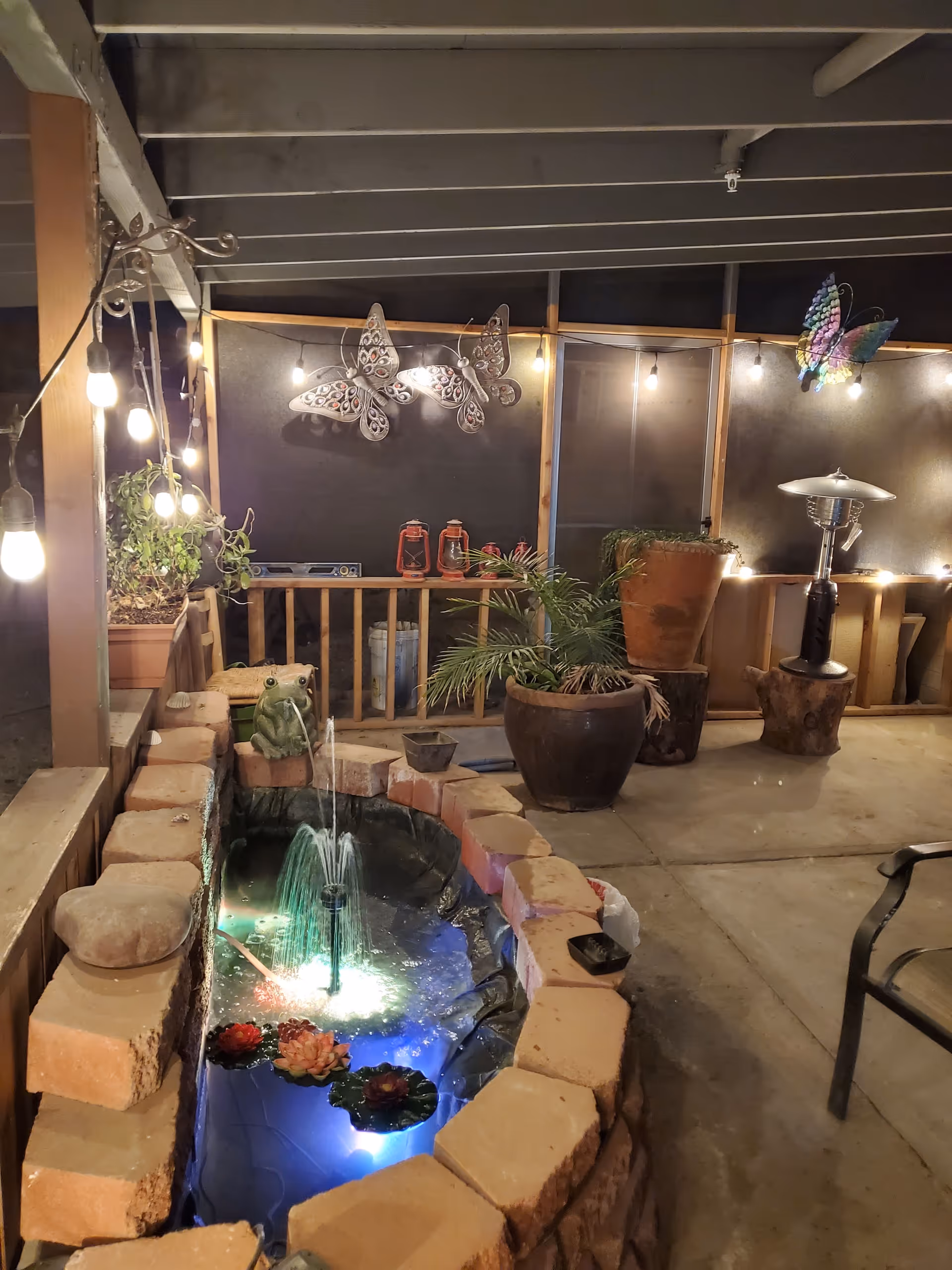 Covered outdoor patio area at night with string lights hanging from the ceiling. A small brick-lined water fountain with a frog statue and floating flowers is illuminated with underwater lights. Potted plants and decorative butterfly wall hangings are visible, along with a patio heater and some chairs.