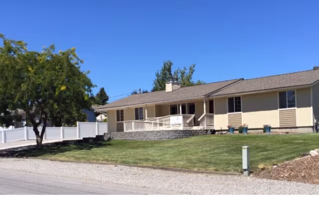 Single-story beige ranch-style home with a front porch, manicured lawn, white fence, and a tree under a clear blue sky.